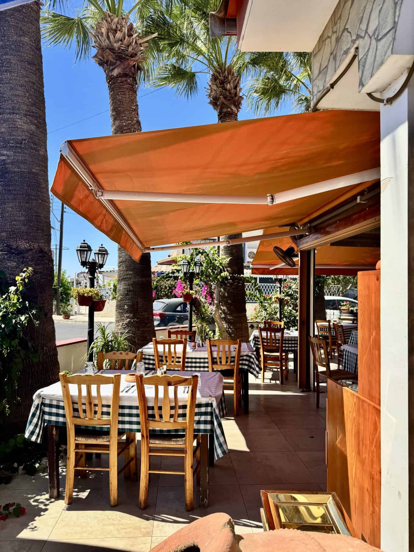 Outdoor dining area of Lysia Meat Tavern with wooden chairs, checkered tablecloths, and orange awning providing shade on a sunny day in Larnaca.