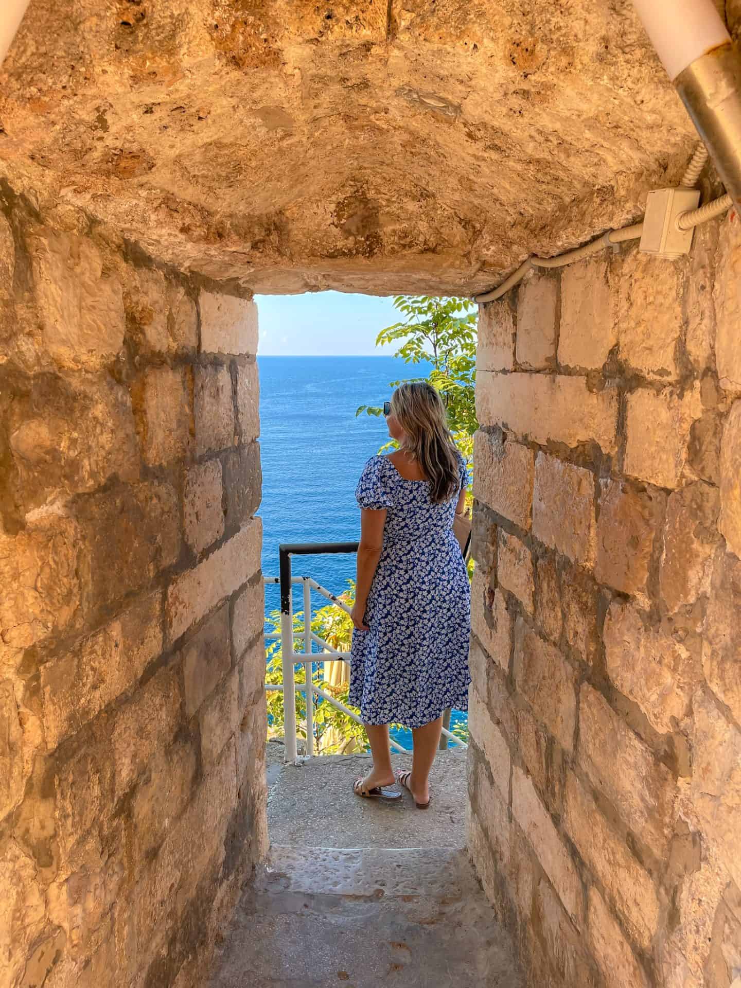 woman in blue dress standing in Stone doorway entrance to Buža Bar Dubrovnik along Old Town walls