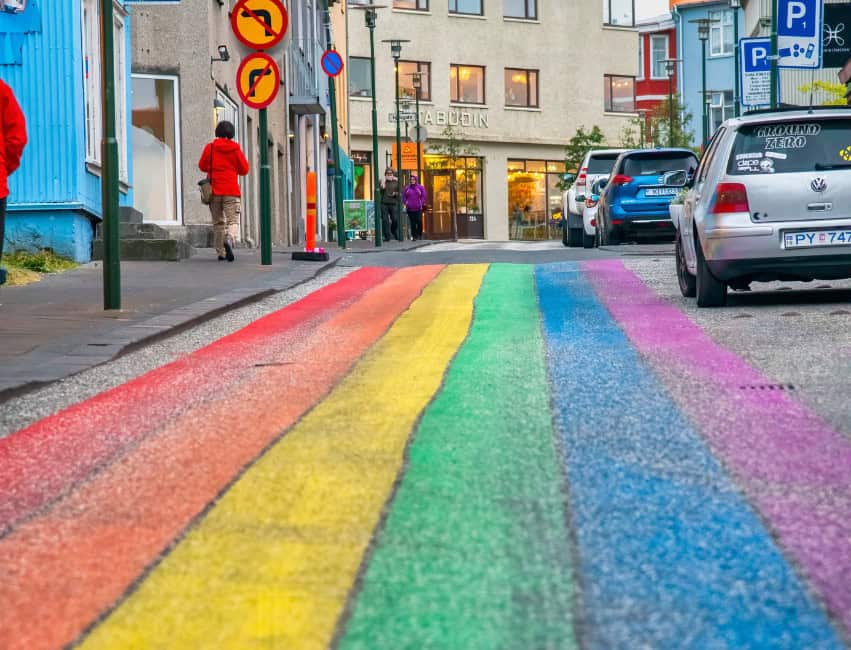 woman walking on rainbow painted street with cars parked and buildings