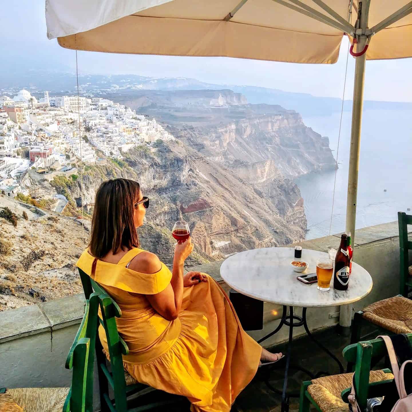 woman in yellow dress sitting with a glass of wine overlooking mountains in fira santorini greece