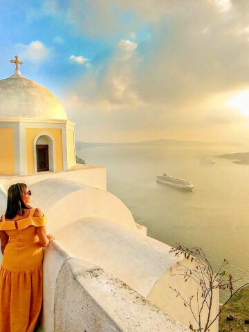 woman in yellow dress looking at sunset with yellow domed church and cruise ship in background in Fira Santorini greece