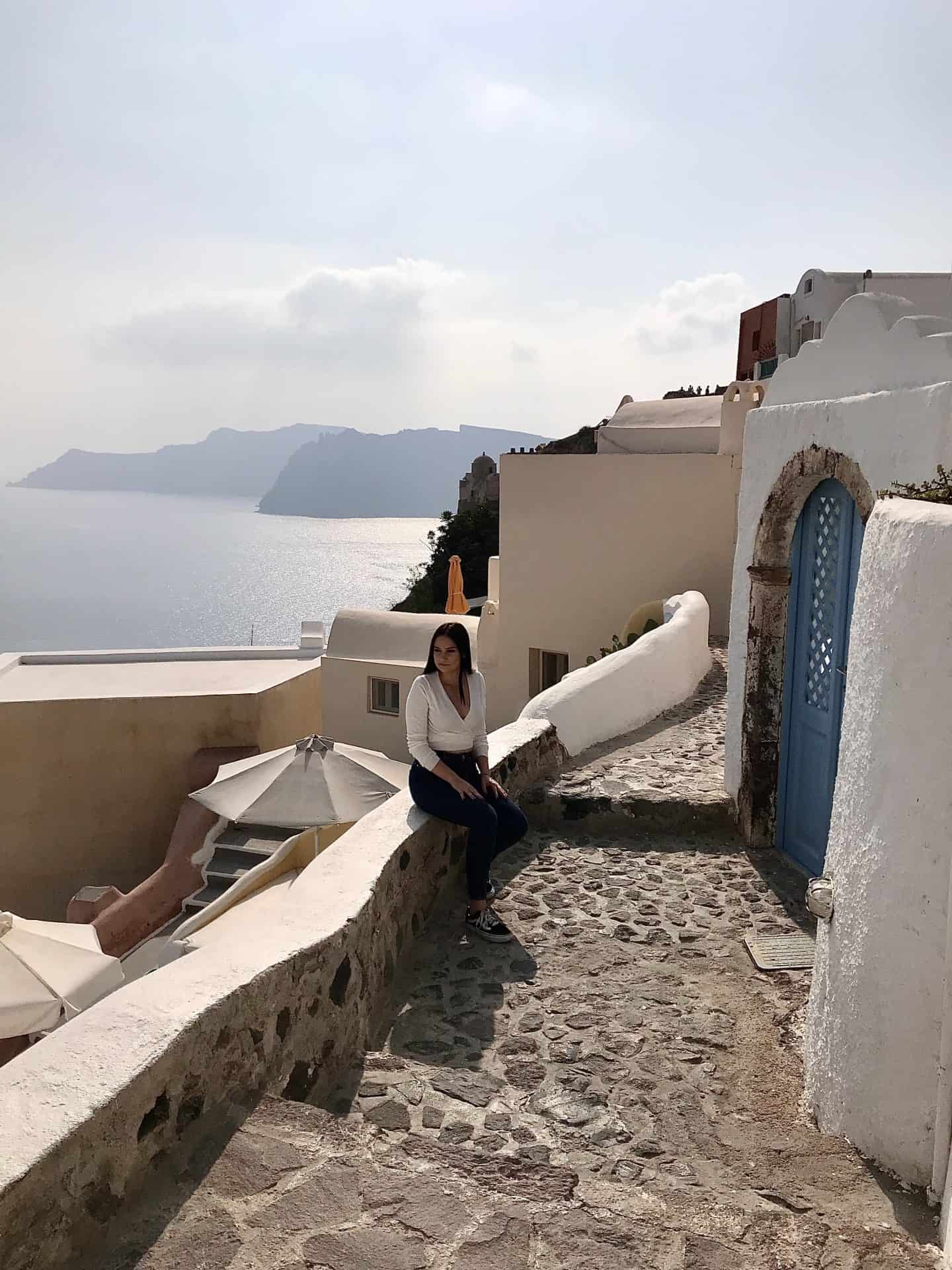 girl in white shirt sitting on stone wall overlooking oia santorini greece